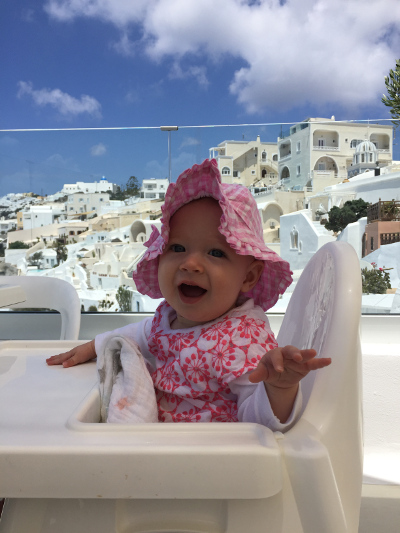 A baby smiles while sitting in a highchair against a backdrop of the whitewashed buildings of Santorini